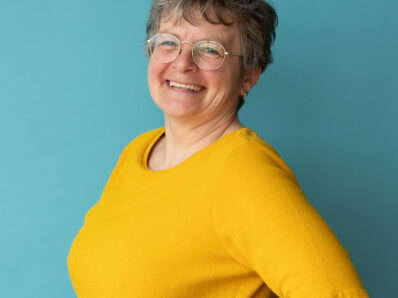 Lachende vrouw met bril, gele trui en hand in haar zij.