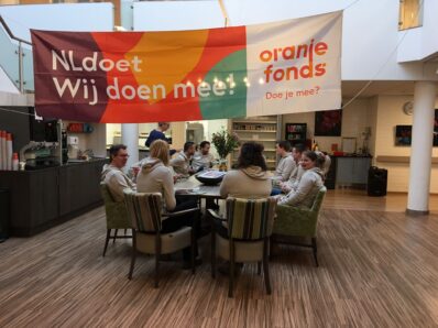 Vrijwilligers zitten aan een tafel, daarboven hangt een vlag van NL Doet